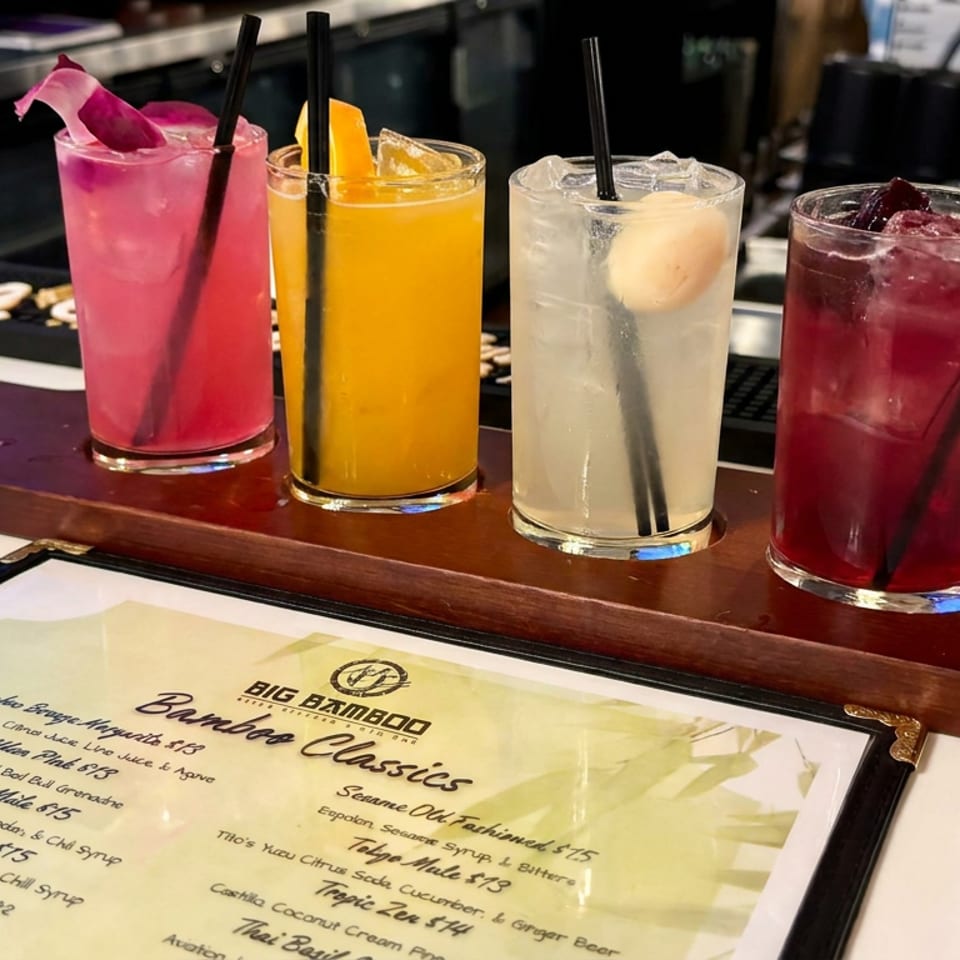 Four colorful cocktails with black straws and fruit garnishes sit on a wooden flight tray beside a Big Bamboo drinks menu.
