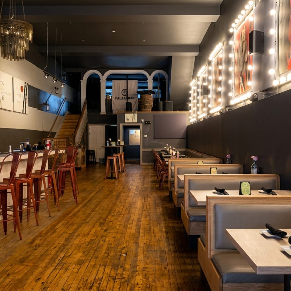 Modern restaurant interior with a long bar, wooden stools, booth seating, polished wood floors, and illuminated wall artwork.