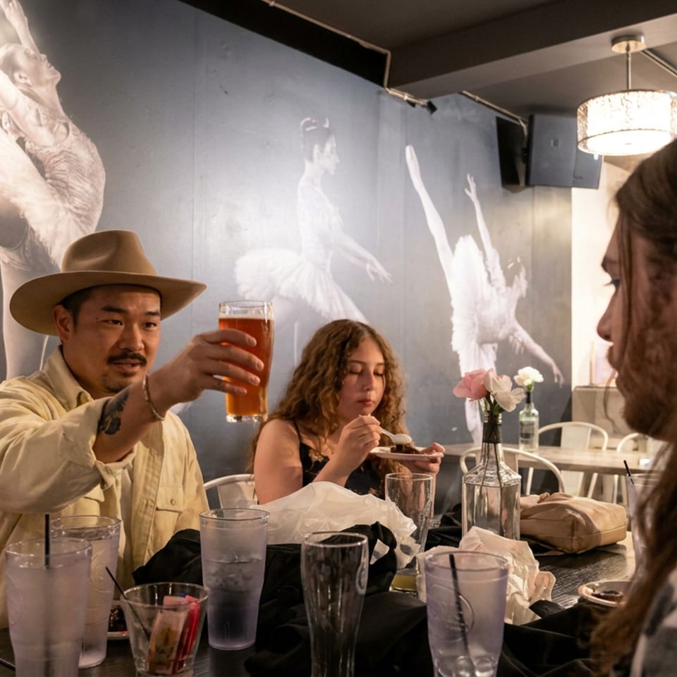 Diners gather around a restaurant table as a man raises a beer, with ballet murals decorating the wall.