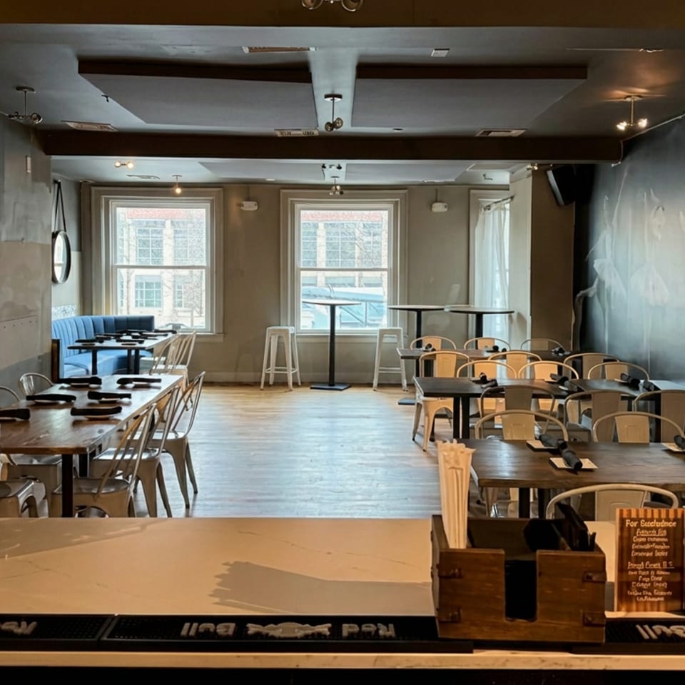 Warmly lit restaurant dining room with neatly set tables, bar counter, large windows, and a ballet-themed wall mural.