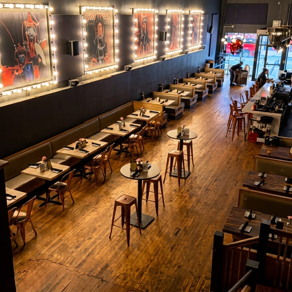Warmly lit restaurant dining room with wooden floors, set tables, booth seating, bar, and colorful artwork in illuminated frames.