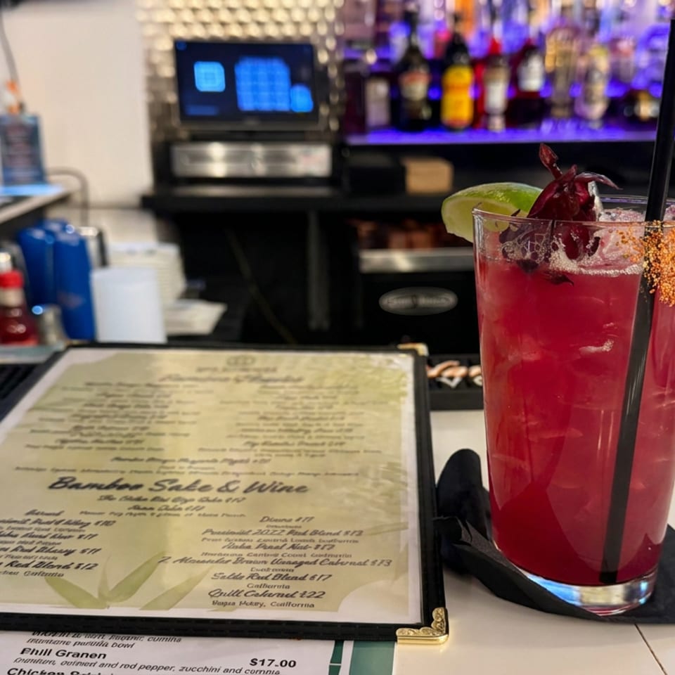 Red cocktail with lime, fruit garnish, and chili-salt rim beside a restaurant menu at a well-stocked bar.