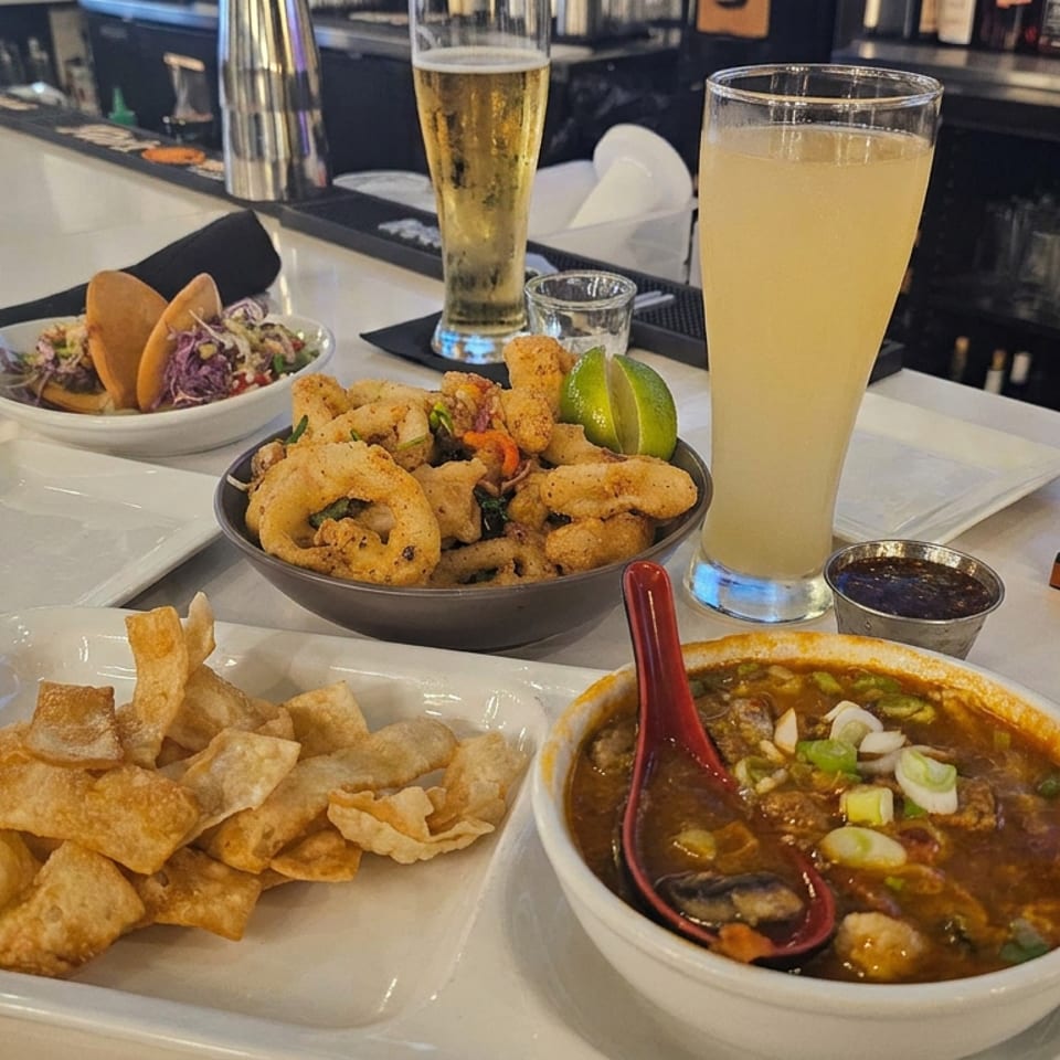 Bar counter spread featuring tacos, fried calamari, crispy wontons, spicy soup, beer, and a pale citrus drink.