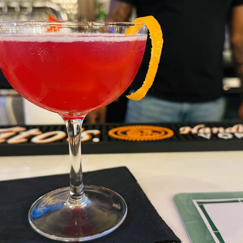 Red cocktail in a coupe glass garnished with an orange peel, resting on a bar counter.