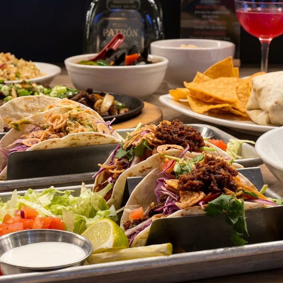 Tex-Mex spread featuring loaded tacos, burrito, tortilla chips, grilled shrimp, vegetables, rice, sauces, and colorful drinks on a restaurant table.