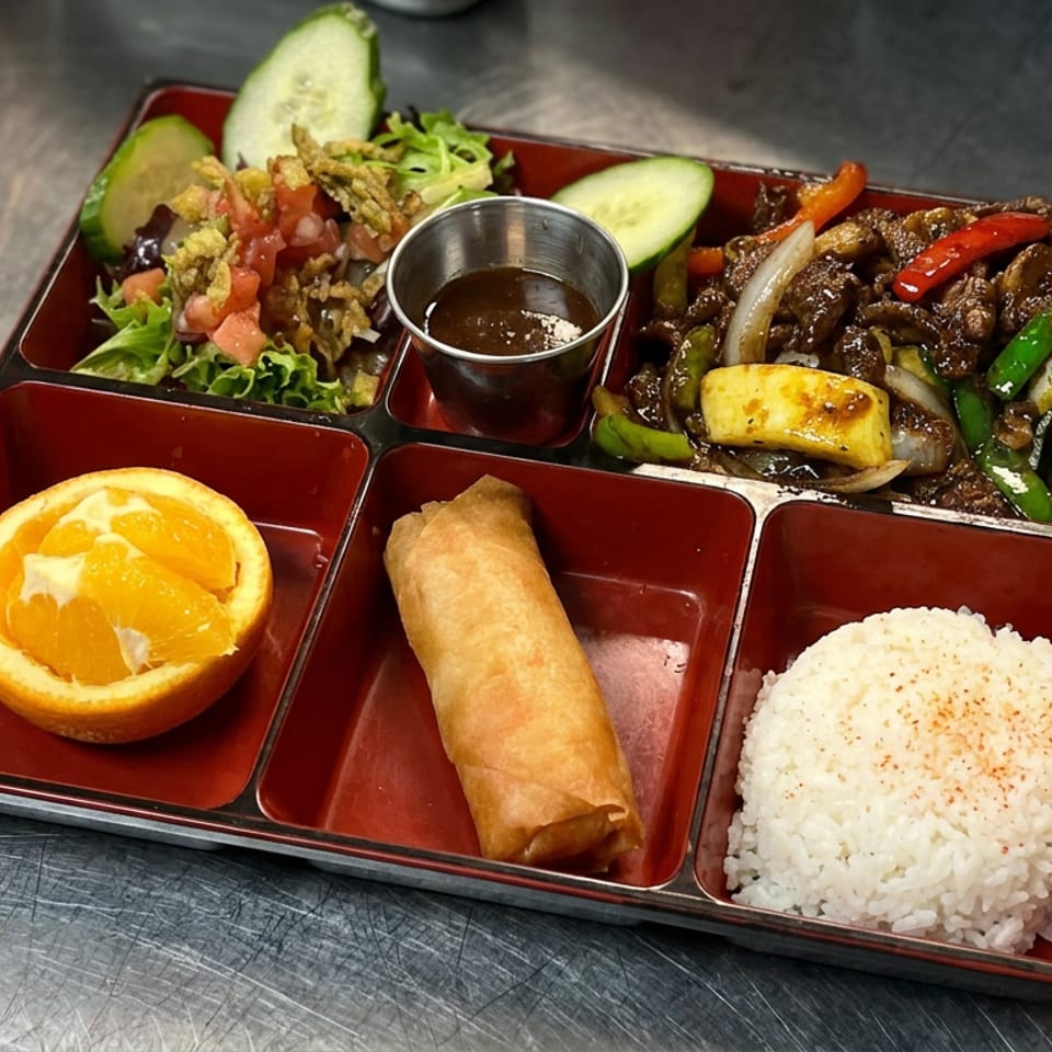 Bento tray with beef stir-fry, salad, spring roll, white rice, orange half, cucumber, and dipping sauce.