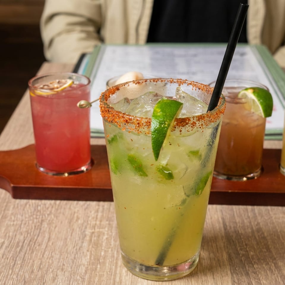 Four colorful cocktails, including a lime-garnished chili-rimmed drink, sit on a wooden serving board at a restaurant table.