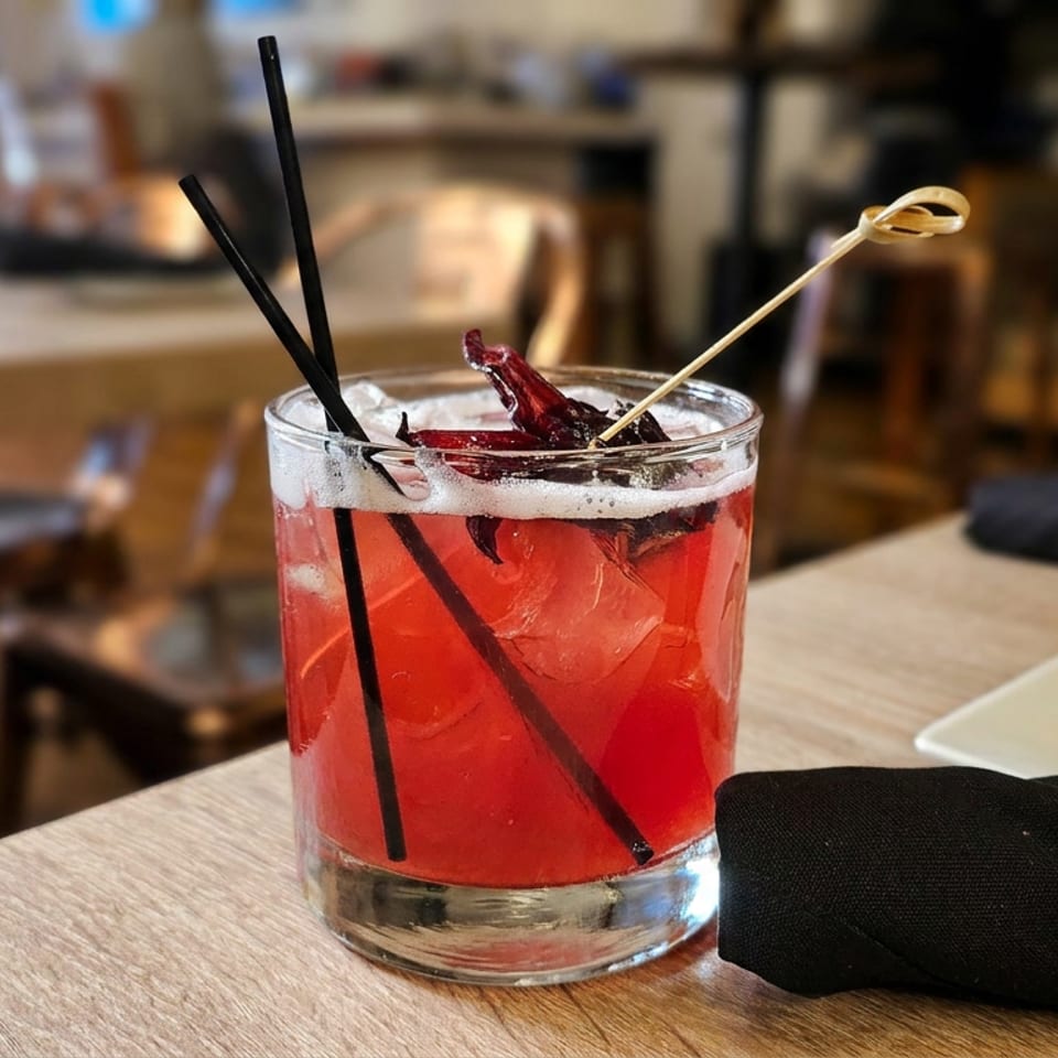 Bright red cocktail with ice, black straws, and a dried red garnish on a restaurant table beside a black napkin.