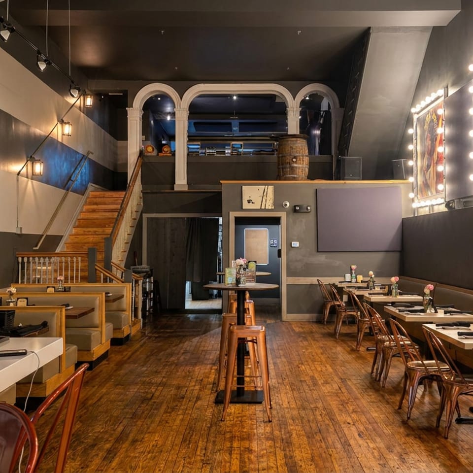 Warmly lit restaurant interior with wood floors, set tables, bar seating, staircase, artwork, and illuminated wall mirrors.